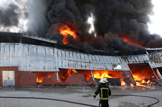 A firefighter directing a hose at an industrial building well alight, flames through the cladding and heavy black smoke