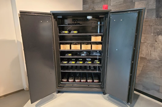 A battery storage cabinet standing open in a workplace, batteries and boxed stock arranged on its internal shelves